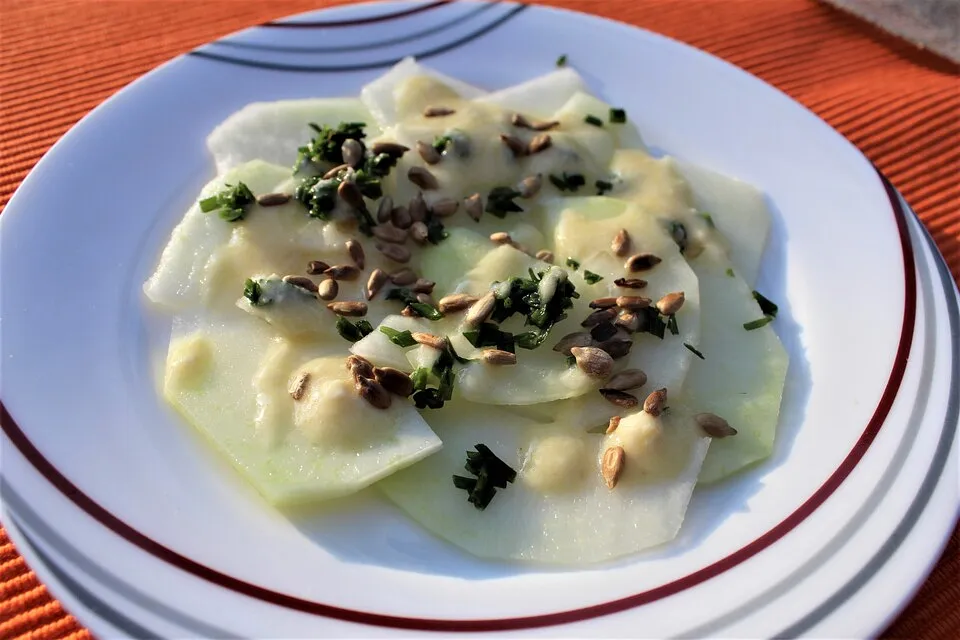 Veganes Kohlrabi-Carpaccio, dünn geschnitten auf einem weißen Teller, garniert mit Sonnenblumenkernen, frischen Kräutern und einer cremigen Sauce.