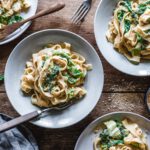 Drei Teller mit veganen Tagliatelle in cremiger Hummus-Wirsing-Sauce, garniert mit gerösteten Kürbiskernen und frischem Wirsing, serviert auf einem rustikalen Holztisch mit einer Schale Gewürzen und einem grauen Leinentuch.