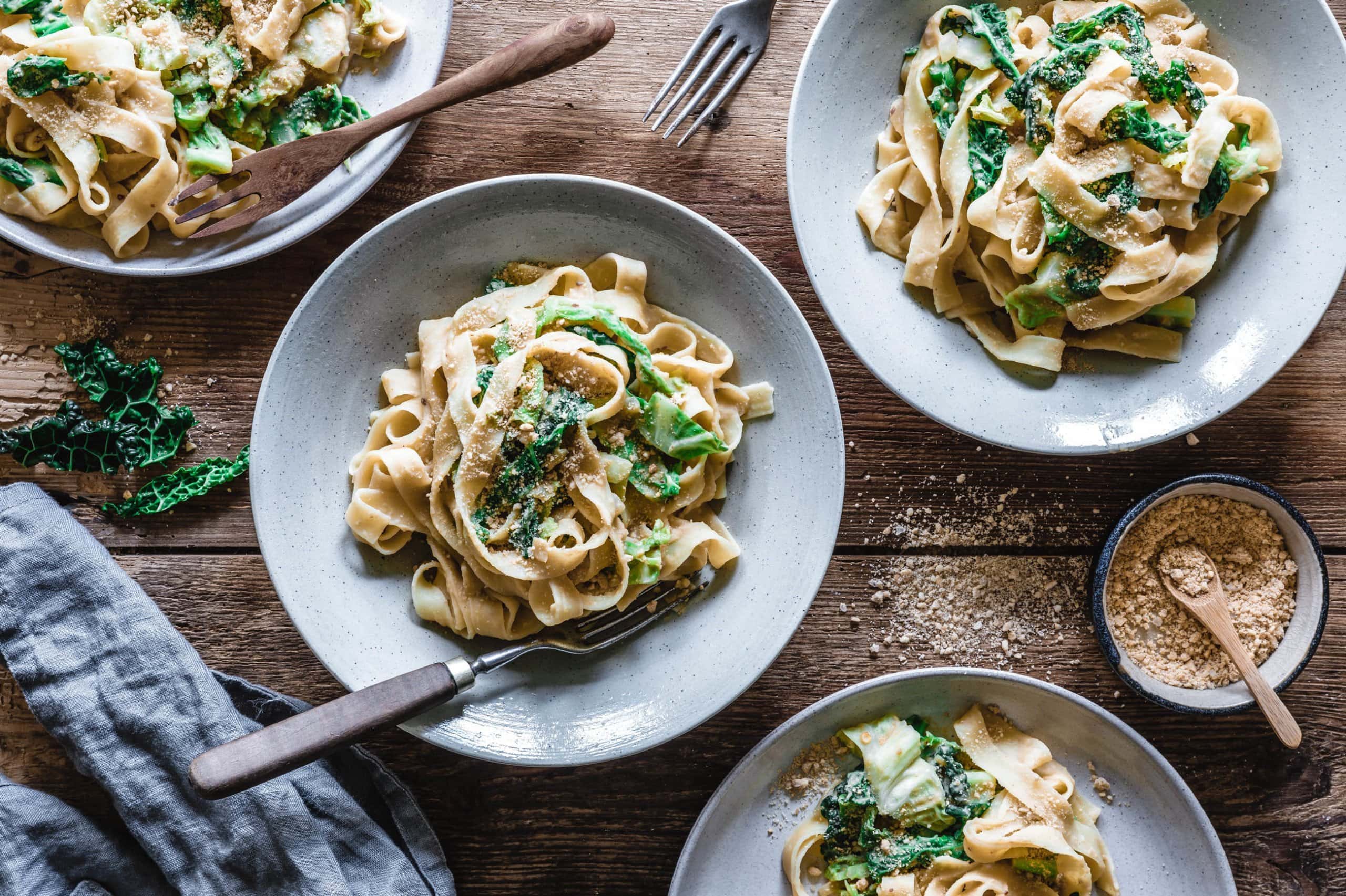 Drei Teller mit veganen Tagliatelle in cremiger Hummus-Wirsing-Sauce, garniert mit gerösteten Kürbiskernen und frischem Wirsing, serviert auf einem rustikalen Holztisch mit einer Schale Gewürzen und einem grauen Leinentuch.