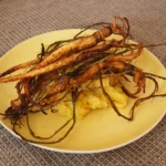 Ein Teller mit gebackener Haferwurzel, die auf einem Bett aus cremigem Süßkartoffelpüree angerichtet ist. Die Haferwurzel ist knusprig gebacken und mit ihren dünnen, leicht verkohlten Wurzelenden dekorativ auf dem Püree drapiert. Der Teller ist gelb und steht auf einer grauen, strukturierten Tischdecke.