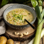 Eine dampfende Schüssel mit veganer Käse-Lauch-Suppe, garniert mit fein geschnittenen Lauchringen. Die Suppe ist cremig und wird in einer grauen Schale serviert, die auf einem rustikalen Holzbrett steht. Um die Schale herum liegen frische Zutaten wie ein ganzer Lauch, Kartoffeln und Cashewkerne, die auf die natürlichen Inhaltsstoffe der Suppe hinweisen. Ein Leinentuch und grüne Blätter im Hintergrund unterstreichen den frischen und gesunden Charakter des Gerichts.