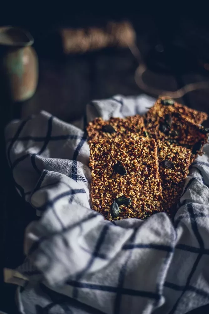 Frisch gebackene Scheiben veganes Roggenvollkorn-Knäckebrot, die in einem karierten Leinentuch liegen. Das Knäckebrot ist goldbraun gebacken und mit verschiedenen Samen wie Kürbiskernen und Sonnenblumenkernen bestreut. Im Hintergrund ist ein dunkler, leicht unscharfer Holztisch zu sehen, der dem Bild eine rustikale und natürliche Atmosphäre verleiht. Das Brot wirkt knusprig und gesund.