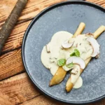 Schwarzwurzeln auf einem dunklen Teller, serviert mit cremiger Hollandaise und in Scheiben geschnittenen Champignons, garniert mit frischem Basilikum.