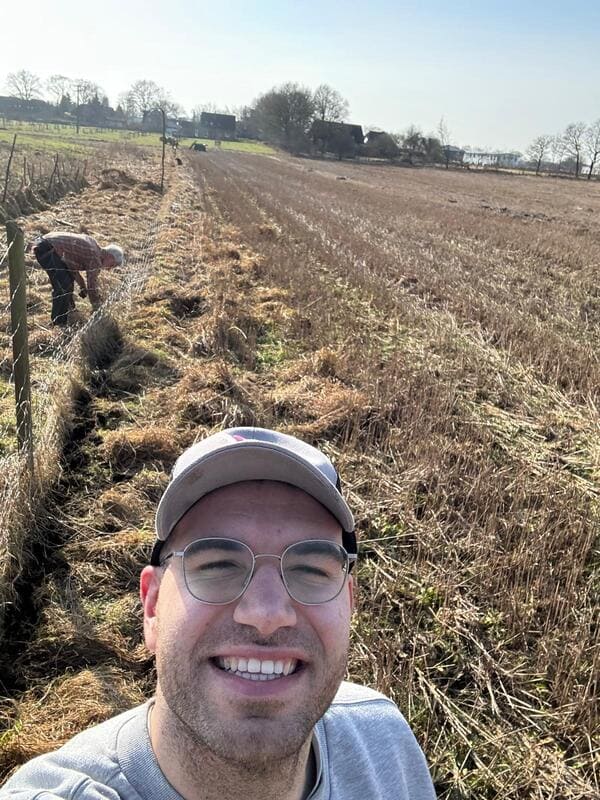 Selfie eines lächelnden Mannes mit Basecap und Brille vor einem Feld, auf dem zwei Personen im Hintergrund arbeiten. Textüberlagerung: "Unsere Arbeit geht weiter!"