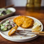 Ein Teller mit knusprigem Kohlrabi-Schnitzel, das goldbraun paniert ist. Dazu wird ein frischer Gurkensalat mit Zwiebeln und einer cremigen Sauce serviert. Eine Zitronenspalte liegt am Rand des Tellers als Garnitur. Im Hintergrund ist ein weiterer Teller mit Salat zu sehen. Das Essen wird auf einem Holztisch präsentiert, und eine Gabel liegt auf dem Teller. Das Bild vermittelt einen appetitlichen und frischen Eindruck.