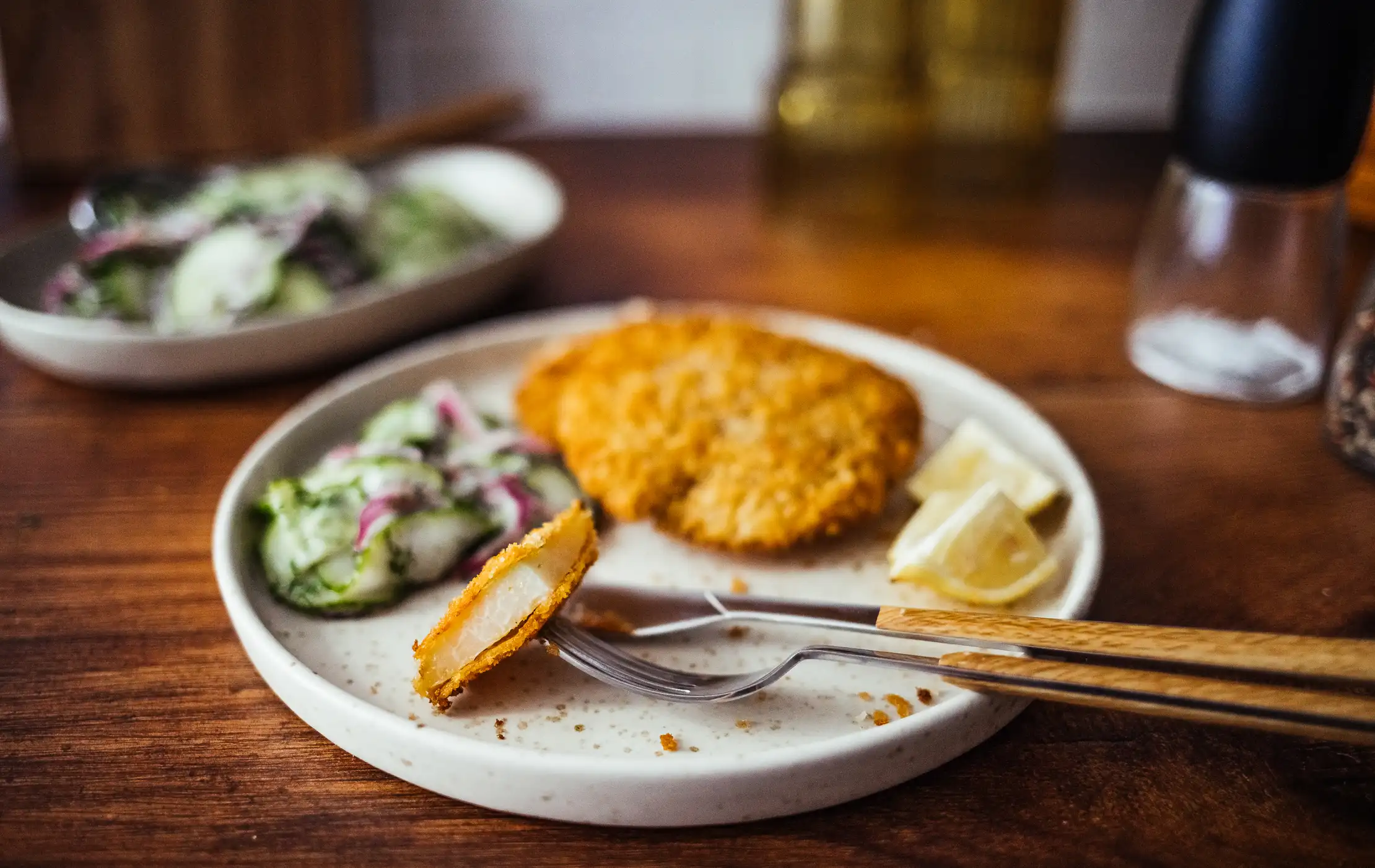 Ein Teller mit knusprigem Kohlrabi-Schnitzel, das goldbraun paniert ist. Dazu wird ein frischer Gurkensalat mit Zwiebeln und einer cremigen Sauce serviert. Eine Zitronenspalte liegt am Rand des Tellers als Garnitur. Im Hintergrund ist ein weiterer Teller mit Salat zu sehen. Das Essen wird auf einem Holztisch präsentiert, und eine Gabel liegt auf dem Teller. Das Bild vermittelt einen appetitlichen und frischen Eindruck.