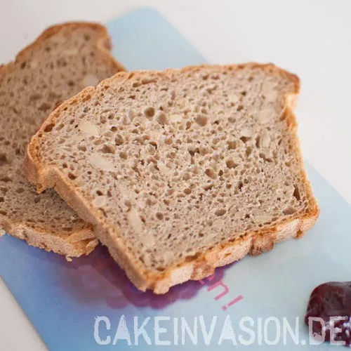 Eine Nahaufnahme von zwei Scheiben frischem Roggenbrot, die leicht versetzt auf einem hellblauen Untergrund liegen. Das Brot hat eine feinporige, bräunliche Krume mit sichtbaren Kernen oder Getreidestückchen und eine knusprige Kruste. In der unteren rechten Ecke ist ein kleiner Klecks dunkle Marmelade zu sehen. Am unteren Bildrand befindet sich der weiße Schriftzug „CAKEINVASION.DE“.