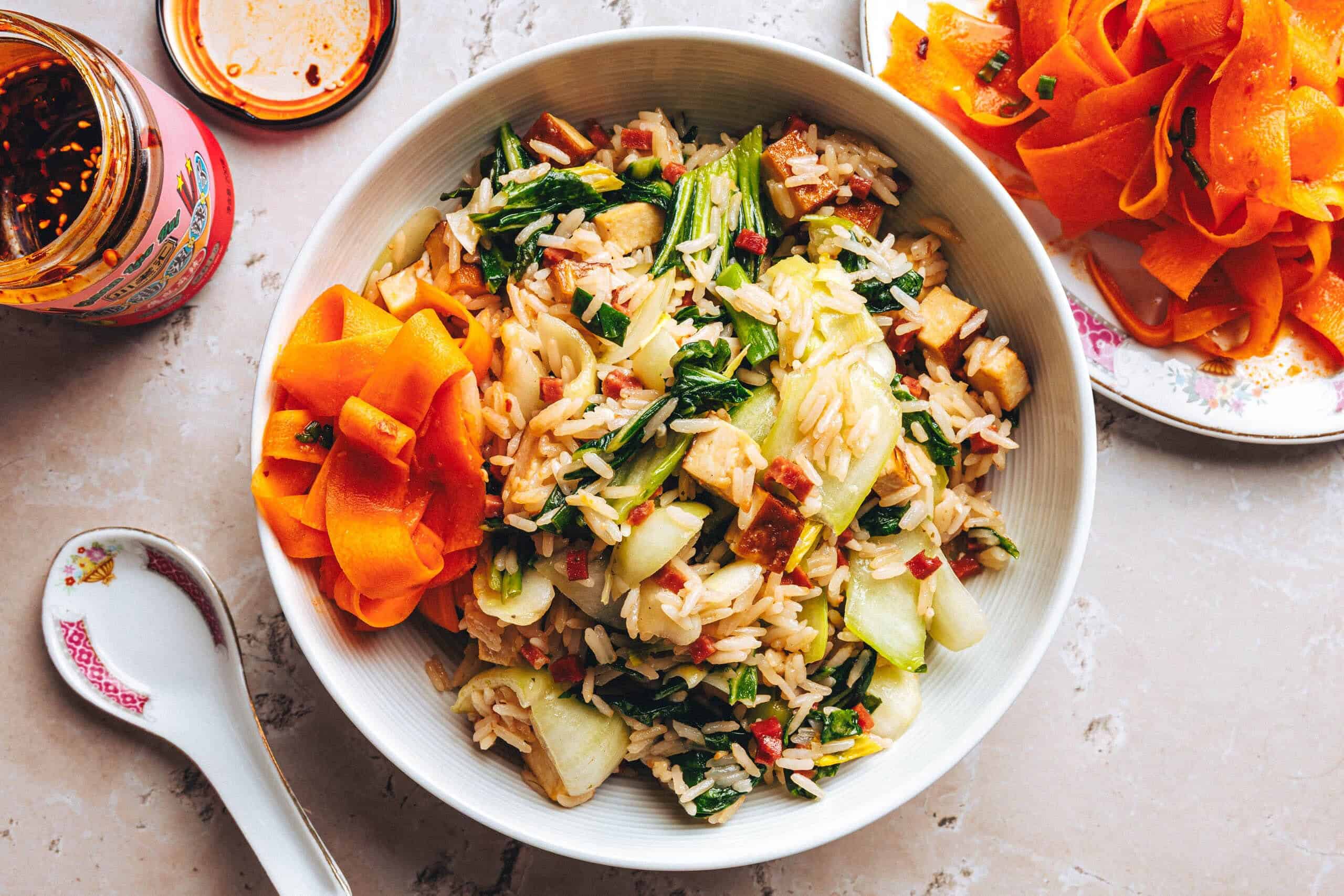 Eine helle Keramikschale, gefüllt mit gebratenem Reis, Pak Choi und goldbraun angebratenen Tofuwürfeln. Garniert ist der Reis mit leuchtend orangefarbenen, in Streifen gehobelten Karotten. Neben der Schale liegen ein asiatischer Keramiklöffel, ein Glas mit Chili-Öl sowie ein weiterer kleiner Teller mit marinierten Karottenstreifen auf einer rustikalen Steinfläche. Das Gericht ist farbenfroh und frisch angerichtet.