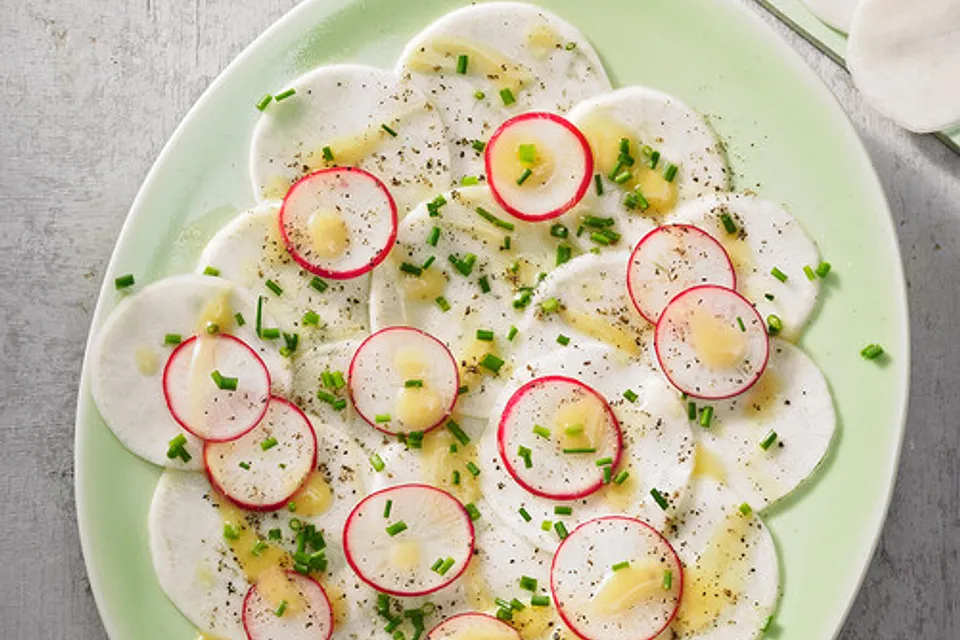 Eine Nahaufnahme von oben auf ein kunstvoll angerichtetes Mairübchen-Carpaccio. Die hauchdünnen, weißen Rübchenscheiben sind fächerförmig auf einem flachen, dunkelgrauen Keramikteller ausgelegt. Das Gericht ist mit frischen, grünen Kräutern, Sprossen, essbaren violetten Blüten und gehackten Nüssen garniert und dezent mit Olivenöl beträufelt. Der Teller steht auf einer rustikalen Holzoberfläche.
