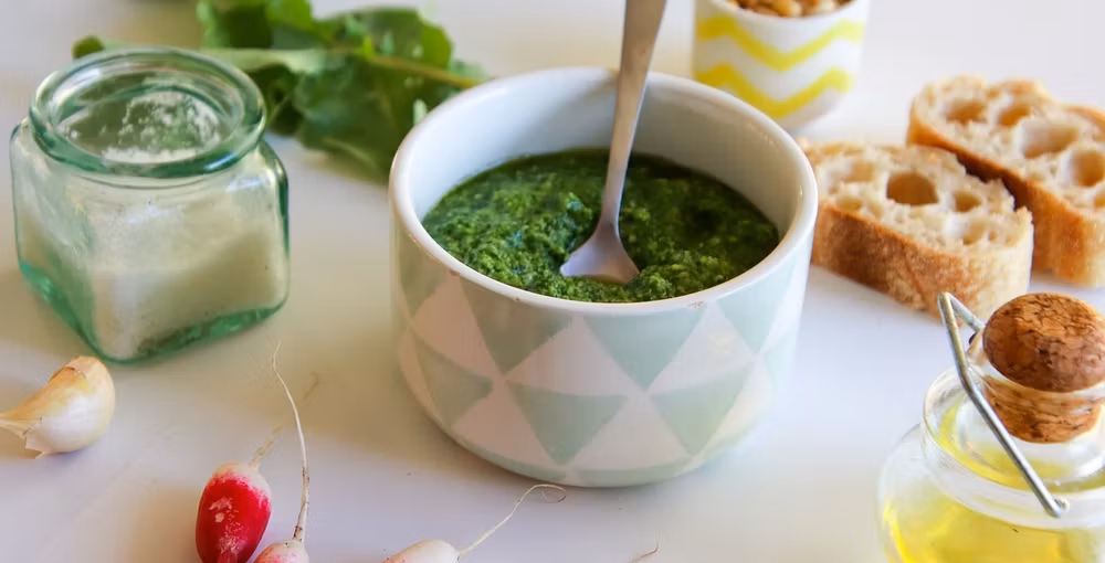 Eine helle Nahaufnahme einer Schale mit frischem grünem Pesto, in der ein kleiner Löffel steckt. Die Schale hat ein dezentes Rautenmuster in Pastelltönen. Um die Schale herum sind Zutaten wie Radieschen, eine Knoblauchzehe, ein Glas mit weißem Pulver (evtl. Salz oder Parmesan), eine Flasche Olivenöl und aufgeschnittenes Baguette dekorativ auf einer weißen Oberfläche arrangiert.
